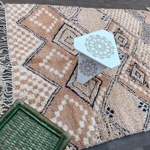 May include: A beige and cream geometric patterned rug with a white, three-legged table. The rug features diamond and checkered patterns with black accents. A green tray is in the foreground.