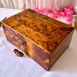 May include: A wooden box with a heart-shaped keyhole and a decorative key. The box is made of a light brown wood with a dark brown wood trim. The box is closed and has a glossy finish.