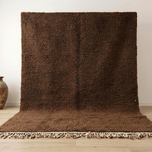 May include: A large, rectangular brown rug with a shaggy texture. The rug is displayed against a white wall, with a portion of it extending onto a wooden floor. The rug has a fringe along the bottom edge. A decorative vase is visible on the left.