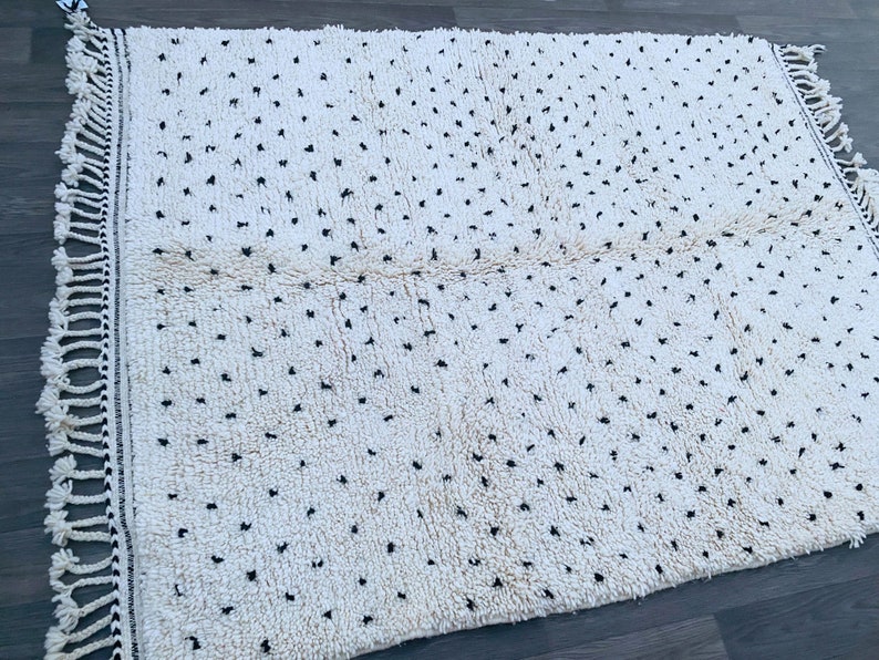 May include: A white rug with black dots and fringe on all sides. The rug has a textured, fluffy surface.