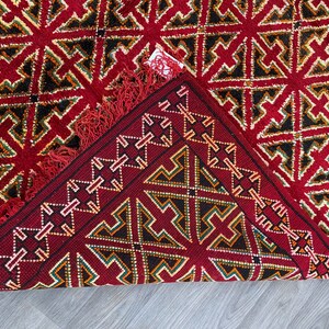 K&ouml;nnte beinhalten: Ein roter Teppich mit einem geometrischen Muster aus schwarzen, gr&uuml;nen und wei&szlig;en Dreiecken. Der Teppich hat einen Fransenrand und ist in der Mitte gefaltet.