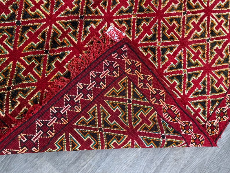 K&ouml;nnte beinhalten: Ein roter Teppich mit einem geometrischen Muster aus schwarzen, gr&uuml;nen und wei&szlig;en Dreiecken. Der Teppich hat einen Fransenrand und ist in der Mitte gefaltet.