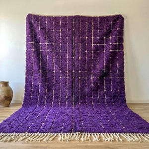 May include: A large, rectangular purple rug with a grid pattern and a cream-colored fringe. The rug is made of a thick, textured material and is displayed against a white wall. A decorative vase is in the background.