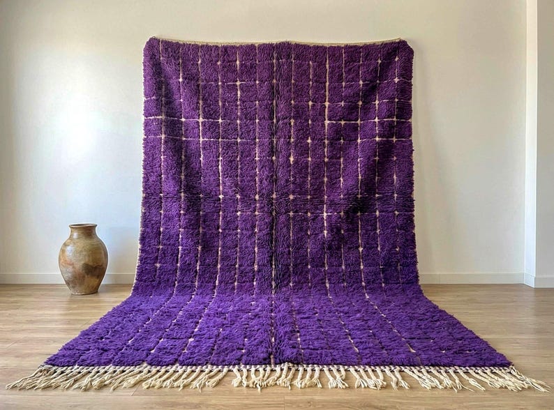 May include: A large, rectangular purple rug with a grid pattern and a cream-colored fringe. The rug is made of a thick, textured material and is displayed against a white wall. A decorative vase is in the background.