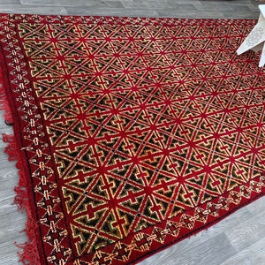 K&ouml;nnte beinhalten: Ein roter, schwarzer und goldener geometrisch gemusterter Teppich mit Fransen. Der Teppich hat ein sich wiederholendes Muster aus Sternen und Kreuzen.