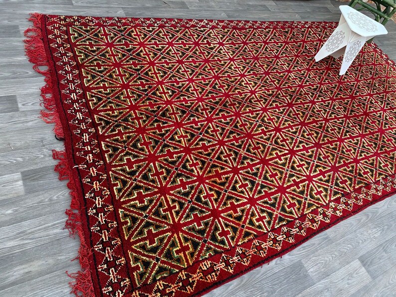 K&ouml;nnte beinhalten: Ein roter, schwarzer und goldener geometrisch gemusterter Teppich mit Fransen. Der Teppich hat ein sich wiederholendes Muster aus Sternen und Kreuzen.