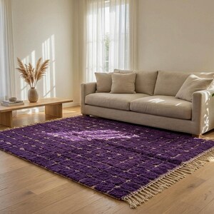 May include: A vibrant purple area rug with a geometric pattern and a fringed edge. The rug is placed on a hardwood floor in a living room setting, with a beige sofa and natural light.