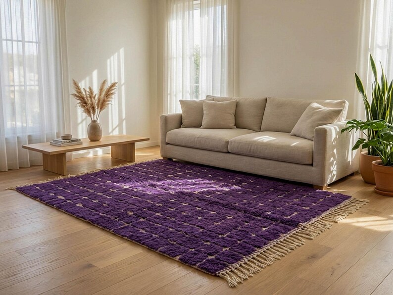 May include: A vibrant purple area rug with a geometric pattern and a fringed edge. The rug is placed on a hardwood floor in a living room setting, with a beige sofa and natural light.