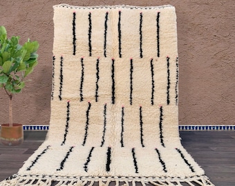 May include: Cream-colored rug with vertical black stripes and fringe. The rug has a textured, woven appearance. Small pink accents are visible between some of the black stripes. The rug is displayed on a wooden floor.