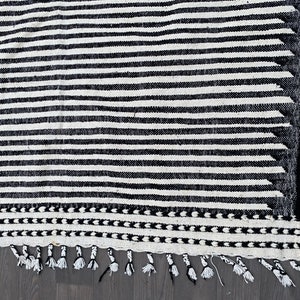 Moroccan handmade rug featuring a striped design in alternating black and white tones (see photos). Crafted from natural wool, this Berber carpet blends durability with elegance. The fringed edges add cultural detail, making it a versatile choice for contemporary living rooms, Scandinavian interiors, or bohemian spaces seeking artisanal charm and timeless heritage.