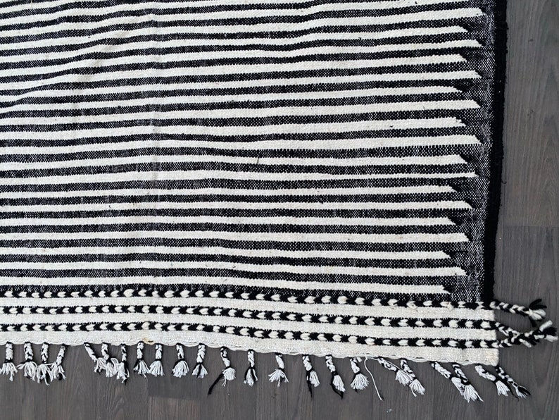 Moroccan handmade rug featuring a striped design in alternating black and white tones (see photos). Crafted from natural wool, this Berber carpet blends durability with elegance. The fringed edges add cultural detail, making it a versatile choice for contemporary living rooms, Scandinavian interiors, or bohemian spaces seeking artisanal charm and timeless heritage.