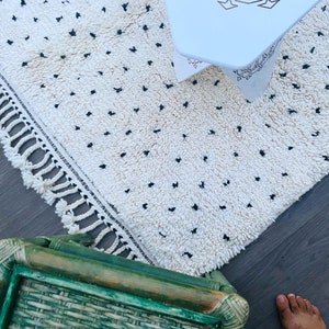 May include: A white rug with black dots and fringe along the edges. The rug is on a wooden floor with a green wicker chair and a white table in the background.