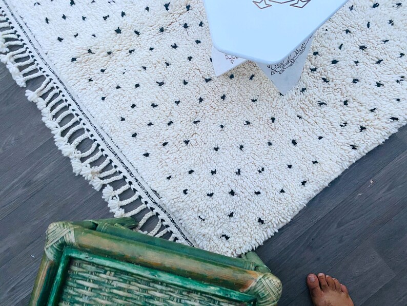 May include: A white rug with black dots and fringe along the edges. The rug is on a wooden floor with a green wicker chair and a white table in the background.