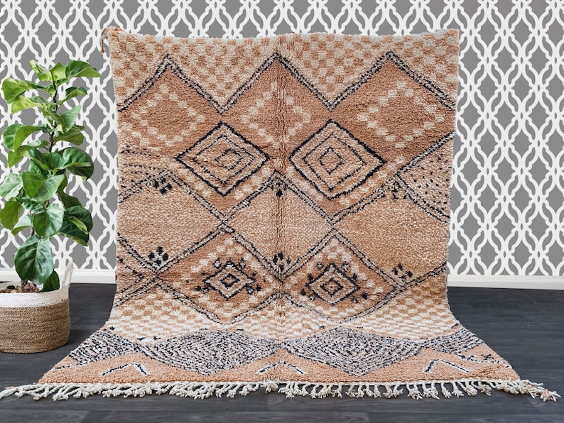 May include: A beige and brown geometric patterned rug with a diamond design. The rug has a textured, plush appearance with a fringe along the bottom edge. The design includes black and white accents.