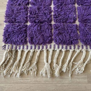 May include: A close-up of a purple and cream-colored rug with a geometric pattern. The rug features a series of square tufted sections in a vibrant purple hue. The bottom edge has cream-colored tassels.