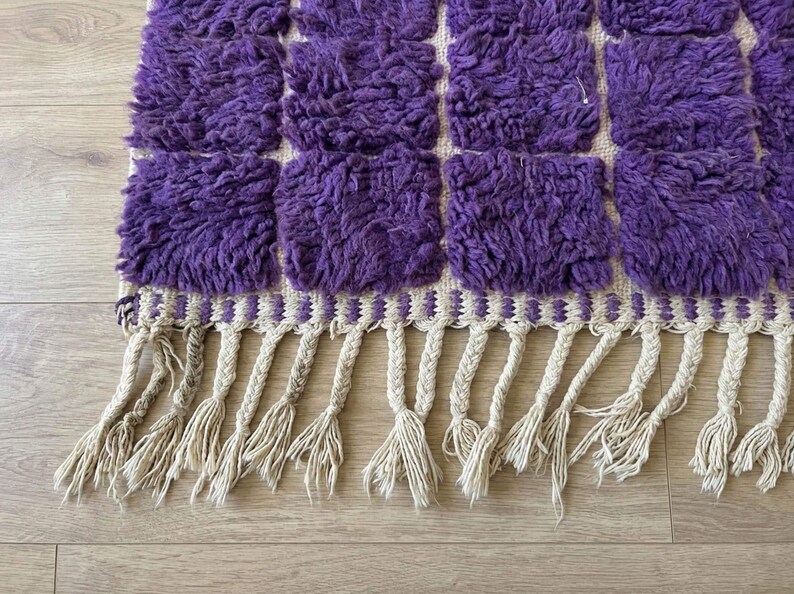 May include: A close-up of a purple and cream-colored rug with a geometric pattern. The rug features a series of square tufted sections in a vibrant purple hue. The bottom edge has cream-colored tassels.