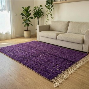 May include: A rectangular purple rug with a textured surface and a fringe border. The rug is placed on a hardwood floor in front of a beige sofa. The room has natural light and indoor plants.