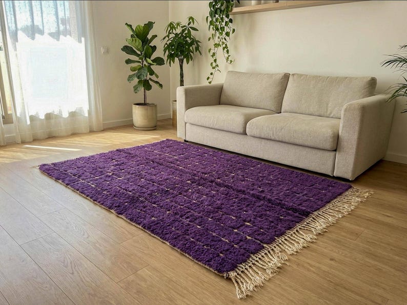 May include: A rectangular purple rug with a textured surface and a fringe border. The rug is placed on a hardwood floor in front of a beige sofa. The room has natural light and indoor plants.
