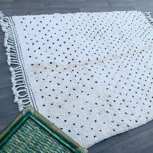 May include: A white rug with a black dot pattern and fringe on all sides. The rug is laid out on a wood floor.