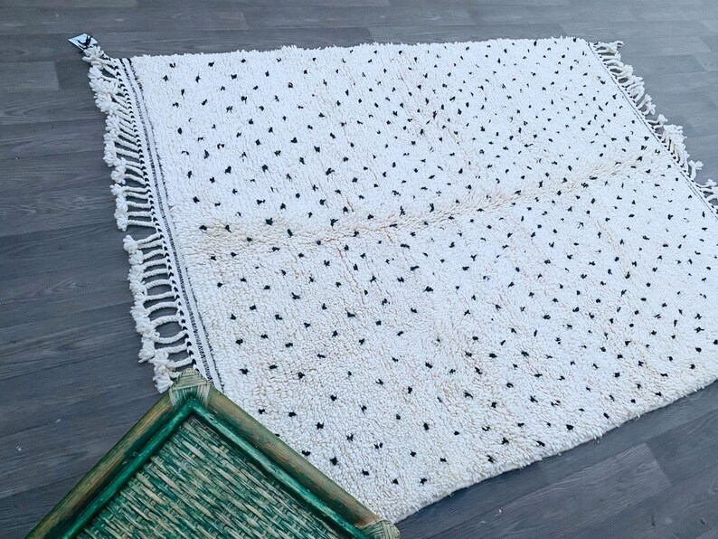 May include: A white rug with a black dot pattern and fringe on all sides. The rug is laid out on a wood floor.