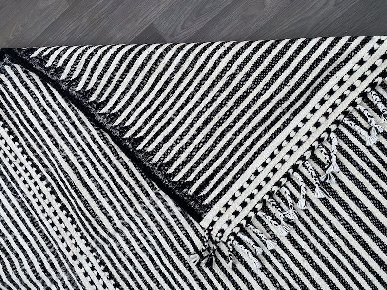 Authentic striped Moroccan rug crafted in ivory and black wool (see photos). Its handwoven linear design highlights Berber craftsmanship with a minimalist twist. Fringes complete the artisanal look, making this rug ideal for boho-chic interiors, Scandinavian homes, or modern spaces seeking cultural depth, comfort, and timeless Moroccan style.