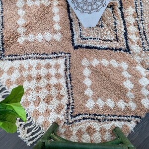 May include: A close-up of a beige and white patterned rug with a geometric design. The rug features a repeating pattern of squares and diamonds, with a fringe along the edge. The rug is made of a soft, textured material.