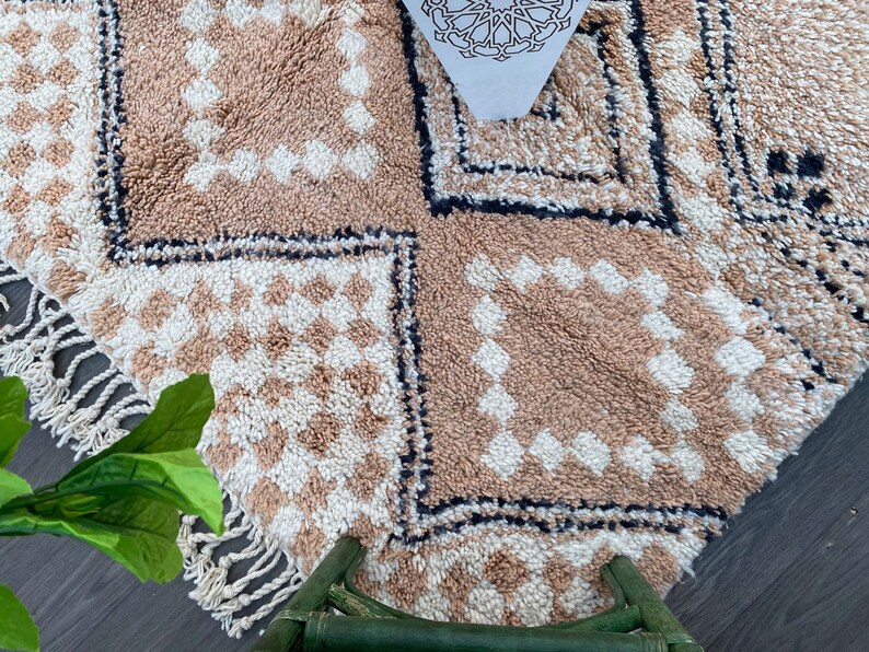 May include: A close-up of a beige and white patterned rug with a geometric design. The rug features a repeating pattern of squares and diamonds, with a fringe along the edge. The rug is made of a soft, textured material.