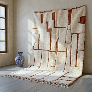 May include: A cream-colored, geometric patterned rug with brown accents hangs against a neutral wall. The rug features a fringe along the bottom edge. A blue and white vase sits near a window.