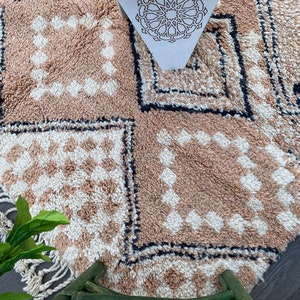 May include: A beige and white patterned rug with a geometric design. The rug features a repeating diamond pattern in beige and white squares, bordered by a black line. The rug has a fringe on one side and a decorative paper with a star pattern.