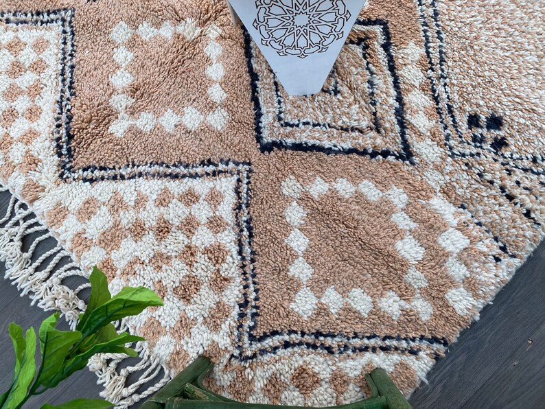 May include: A beige and white patterned rug with a geometric design. The rug features a repeating diamond pattern in beige and white squares, bordered by a black line. The rug has a fringe on one side and a decorative paper with a star pattern.