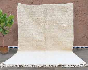 May include: A large, rectangular rug with a cream-colored, textured surface. The rug has a fringe along the bottom edge. The rug is displayed against a brown wall with a small potted plant to the left.