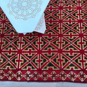 K&ouml;nnte beinhalten: Ein roter Teppich mit einem geometrischen Muster aus schwarzen, wei&szlig;en und goldenen Dreiecken. Der Teppich hat einen Fransenrand.