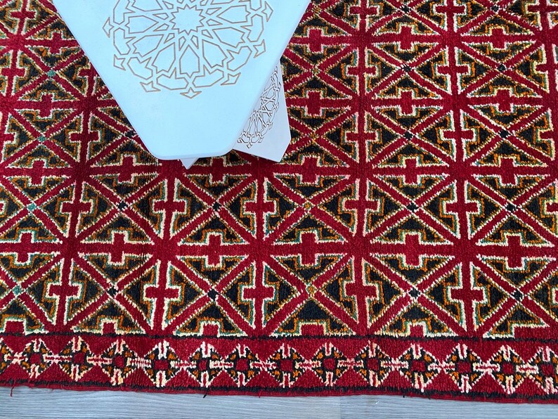 K&ouml;nnte beinhalten: Ein roter Teppich mit einem geometrischen Muster aus schwarzen, wei&szlig;en und goldenen Dreiecken. Der Teppich hat einen Fransenrand.