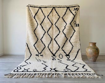 May include: Cream-coloured, hand-woven rug with a geometric diamond pattern in black. The rug has a thick pile, fringe along the bottom edge, and is displayed against a white wall. A decorative, brown ceramic vase is to the right.
