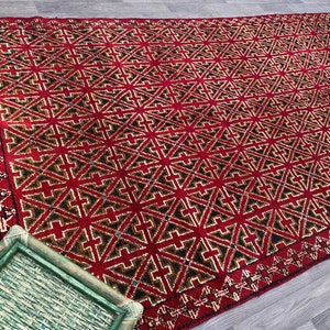 K&ouml;nnte beinhalten: Ein roter Teppich mit einem geometrischen Muster aus schwarzen und goldenen Kreuzen. Der Teppich hat einen Fransenrand.