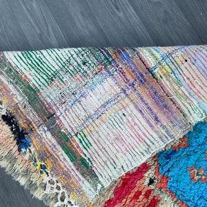 Puede incluir: Una alfombra colorida tejida a mano con un patr&oacute;n geom&eacute;trico en tonos de rosa, azul, verde y blanco. La alfombra tiene un borde de flecos y est&aacute; hecha de lana.
