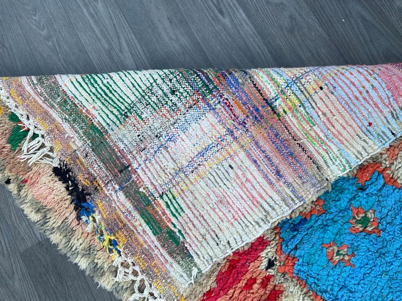 Puede incluir: Una alfombra colorida tejida a mano con un patr&oacute;n geom&eacute;trico en tonos de rosa, azul, verde y blanco. La alfombra tiene un borde de flecos y est&aacute; hecha de lana.