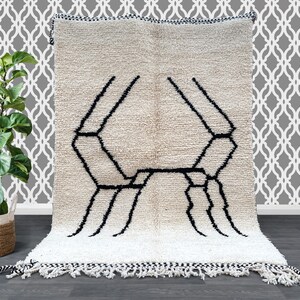 Puede incluir: Una alfombra marroquí blanca y negra con un diseño geométrico. La alfombra tiene un borde de flecos.