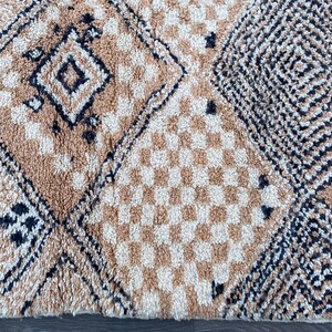 May include: Close-up of a beige and cream-colored rug with geometric patterns. The rug features diamond, square, and checkered designs in varying shades of beige and cream, with black accents. The rug has a textured, plush appearance.