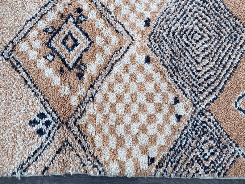 May include: Close-up of a beige and cream-colored rug with geometric patterns. The rug features diamond, square, and checkered designs in varying shades of beige and cream, with black accents. The rug has a textured, plush appearance.