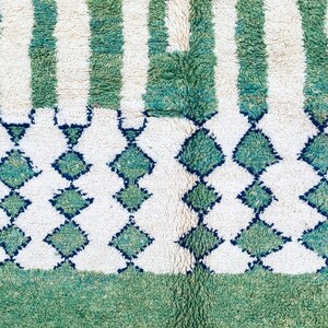K&ouml;nnte beinhalten: Ein wei&szlig;er und gr&uuml;ner Teppich mit geometrischem Muster. Der Teppich hat einen Fransenrand und ist aus weichem, pl&uuml;schigem Material.