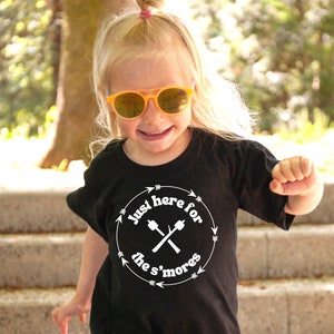 May include: A young child wearing a black t-shirt with the text "Just here for the s'mores" in a white circular design. The child is also wearing orange sunglasses and smiling. The background is a blurred outdoor scene.
