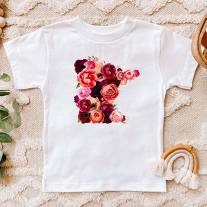May include: White t-shirt featuring a floral design in the shape of the state of Minnesota. The floral arrangement includes red, pink, and purple flowers. The shirt is displayed on a neutral-toned surface.