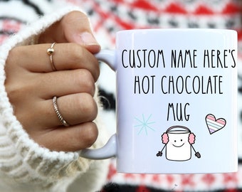 Könnte beinhalten: Weiße Keramik-Tasse mit dem Text "CUSTOM NAME HERE'S HOT CHOCOLATE MUG" und einem Cartoon-Marshmallow-Charakter mit Ohrenschützern. Die Tasse hat auch eine Schneeflocke und ein Herz mit Streifen. Die Tasse wird von einer Person gehalten.