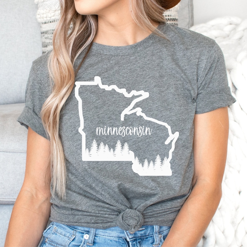 May include: A gray t-shirt featuring a white outline of the states of Minnesota and Wisconsin, with the word "minnesconsin" in a script font. Below the text is a white graphic of a forest. The shirt is tied at the waist.