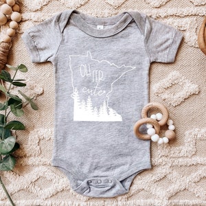 May include: A gray baby onesie with the phrase "Oh Fer Cute" in white, along with a white outline of the state of Minnesota and a forest silhouette. The onesie is surrounded by wooden beads and a green plant.