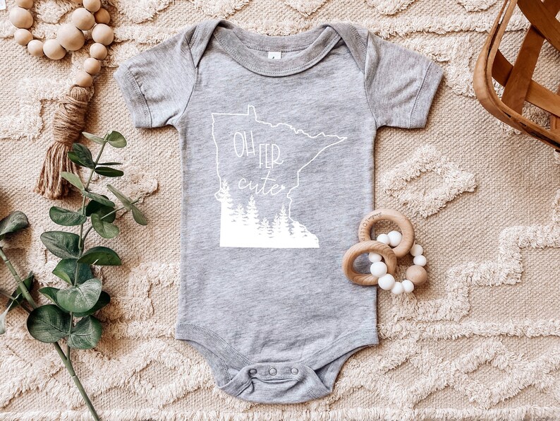 May include: A gray baby onesie with the phrase "Oh Fer Cute" in white, along with a white outline of the state of Minnesota and a forest silhouette. The onesie is surrounded by wooden beads and a green plant.