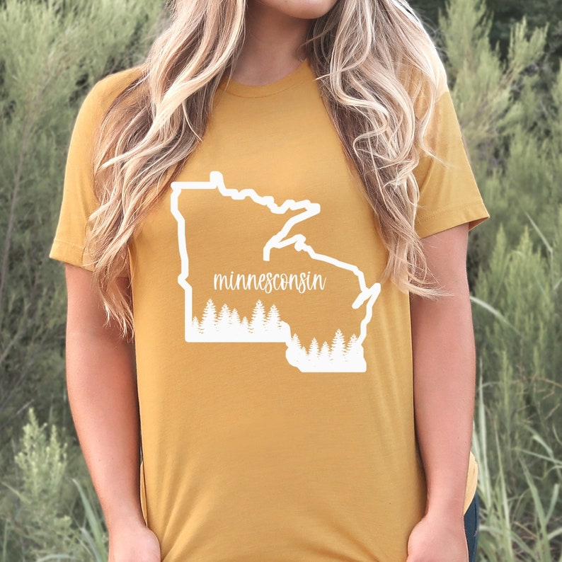 May include: Mustard yellow t-shirt featuring a white outline of the state of Minnesota with the word "minnesconsin" in a script font. Below the state outline is a row of white pine trees. The shirt is made of a soft material.
