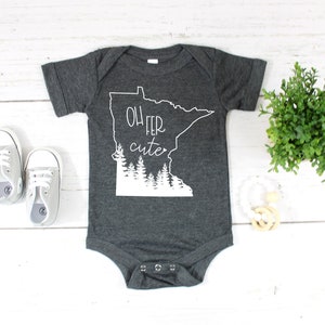 May include: Dark gray baby onesie with the text "Oh fer cute" and a white outline of the state of Minnesota. The onesie also features a white graphic of trees. A pair of gray baby shoes are next to the onesie.