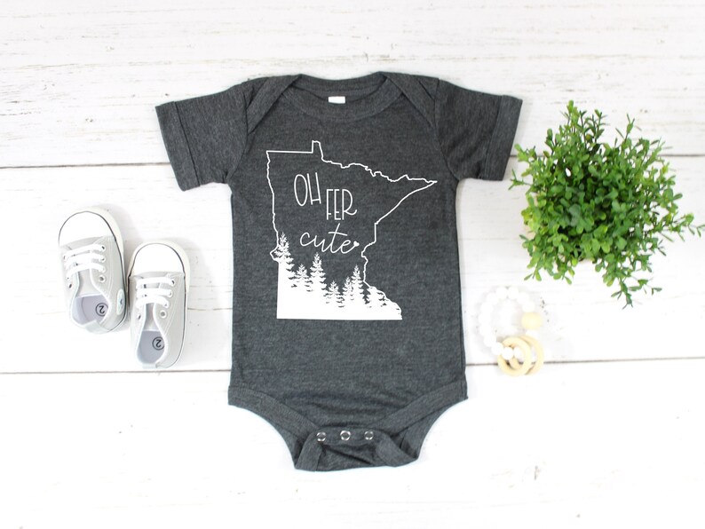 May include: Dark gray baby onesie with the text "Oh fer cute" and a white outline of the state of Minnesota. The onesie also features a white graphic of trees. A pair of gray baby shoes are next to the onesie.